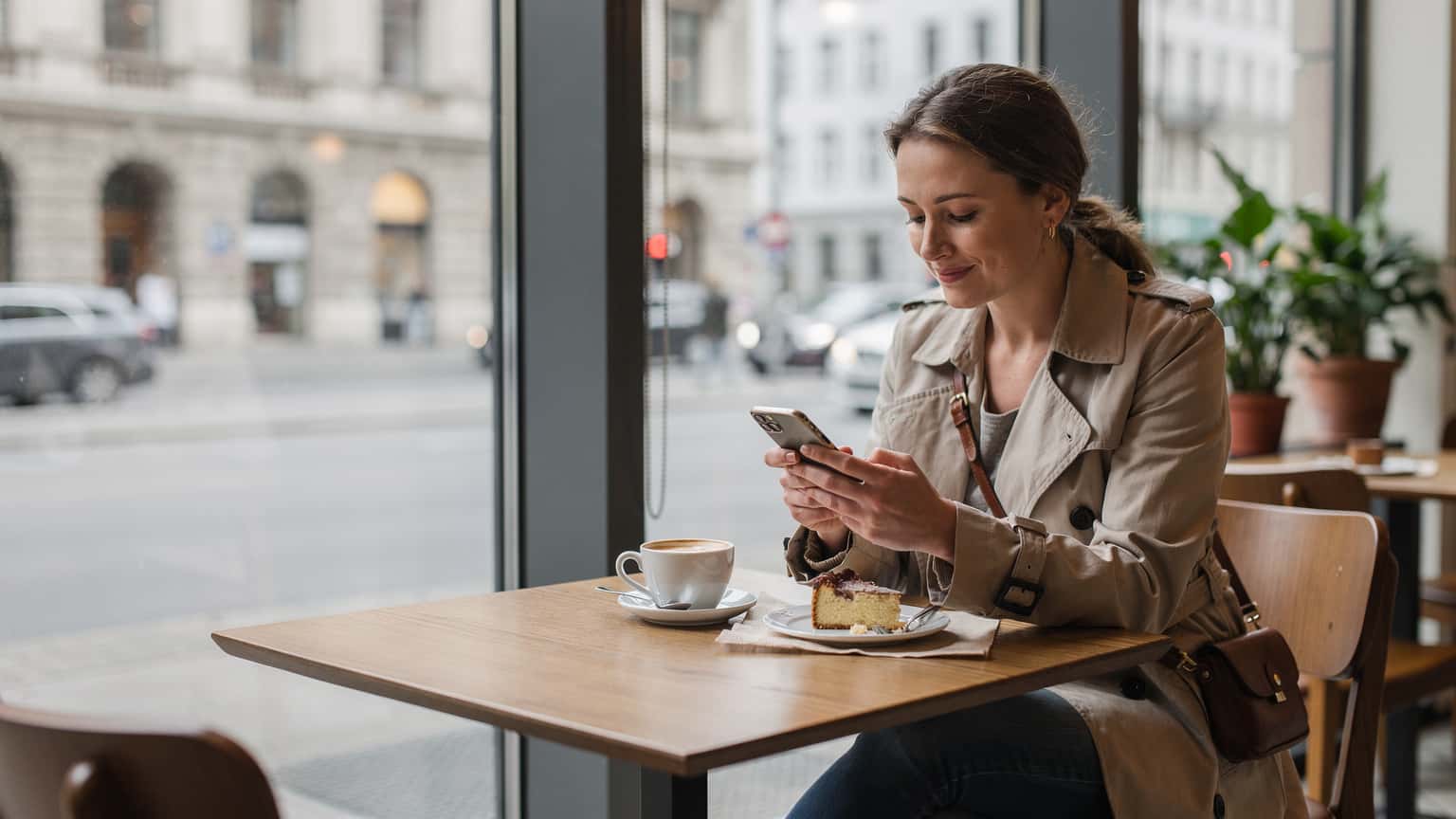Eine Frau sitzt entspannt an einem Fenster in einem modernen Berliner Café, schaut auf ihr Smartphone und genießt einen Kaffee, während sie die Stadtlandschaft überblickt.