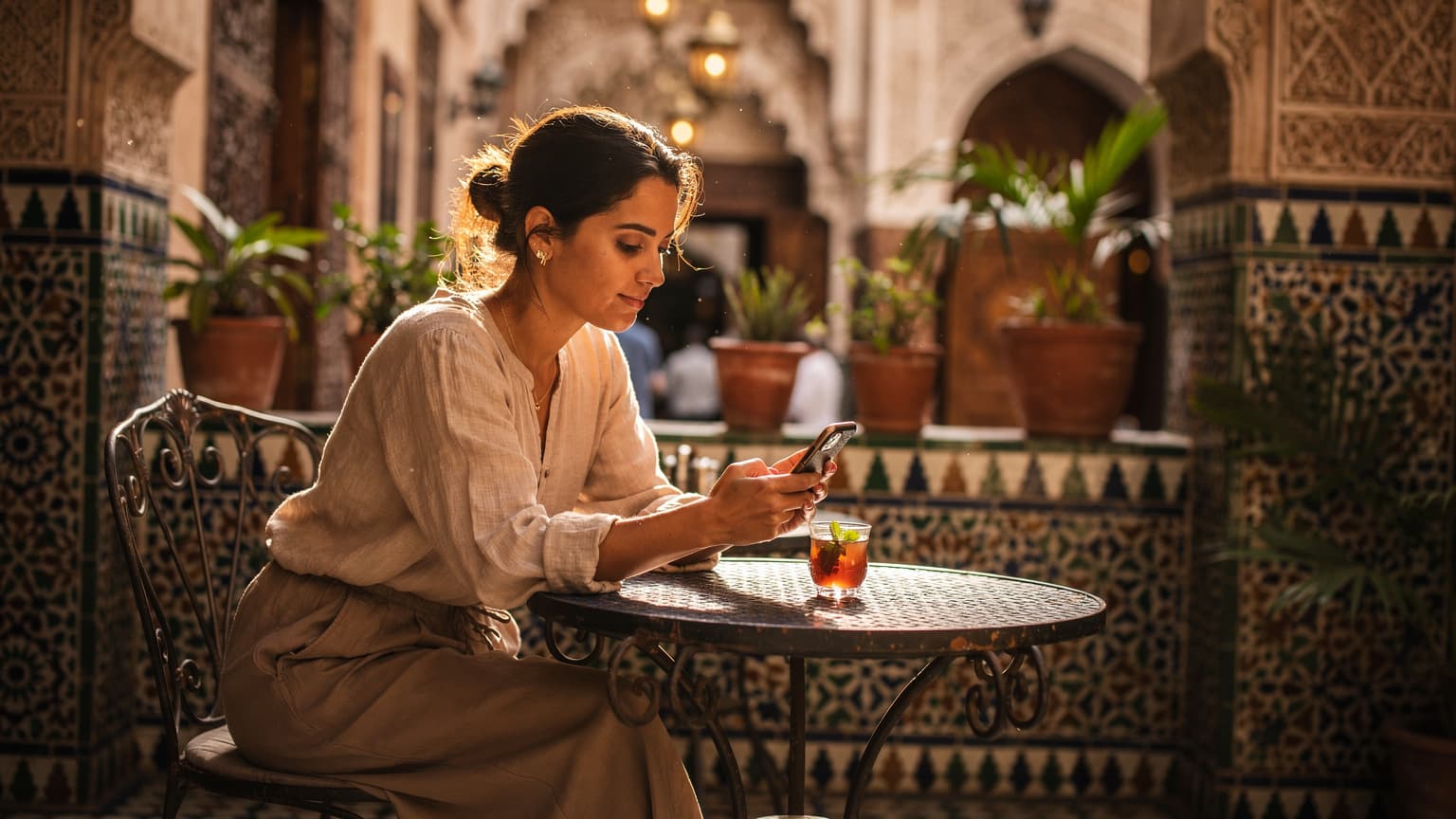 Une femme utilisant son téléphone portable avec une eSIM active, assise à la terrasse d'un café traditionnel à Fès, au Maroc, avec des motifs géométriques en arrière-plan.