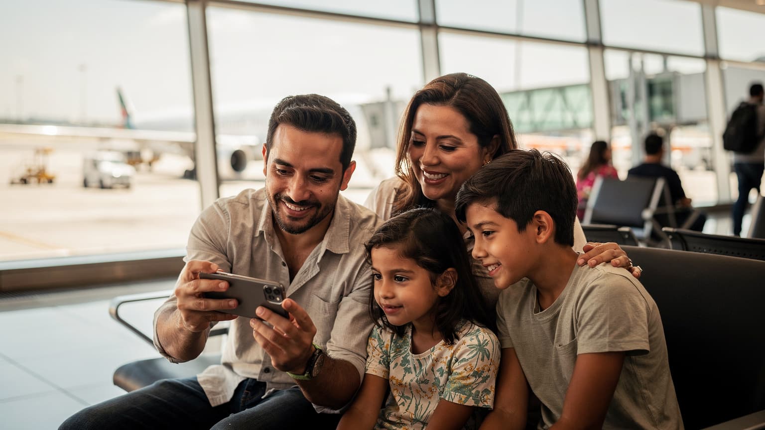 Una familia latina sonriente, con dos niños pequeños y sus padres, interactuando con un teléfono móvil mientras esperan en la sala de embarque de un aeropuerto moderno en Latinoamérica, con grandes ventanales y aviones al fondo.
