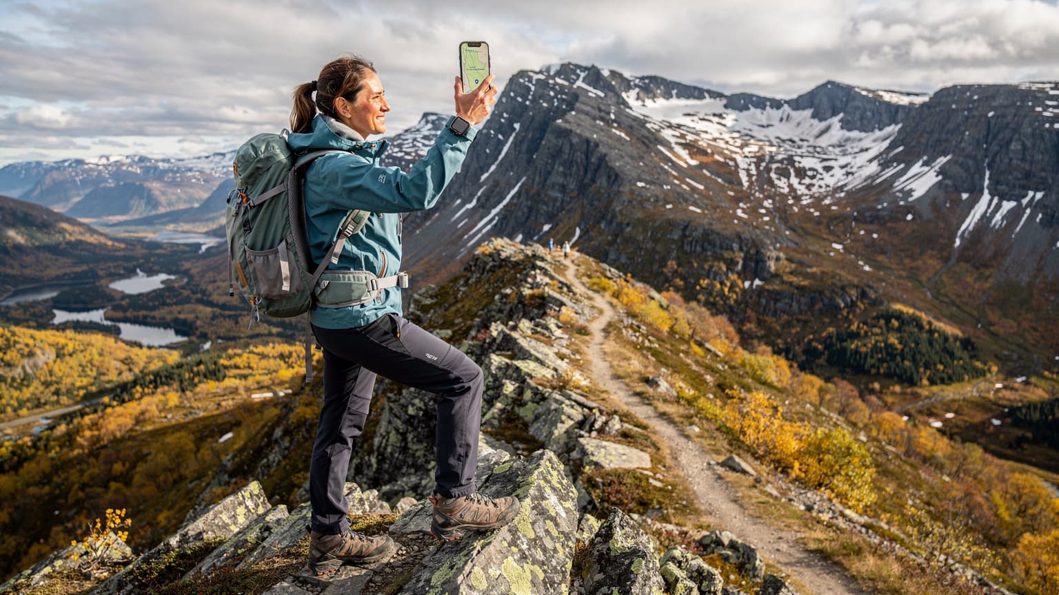 En kvinne i turantrekk står på en fjelltopp i Jotunheimen, ser utover et dramatisk høstlandskap med snødekte topper og gule trær, og holder opp en smarttelefon med en kart-app åpen.