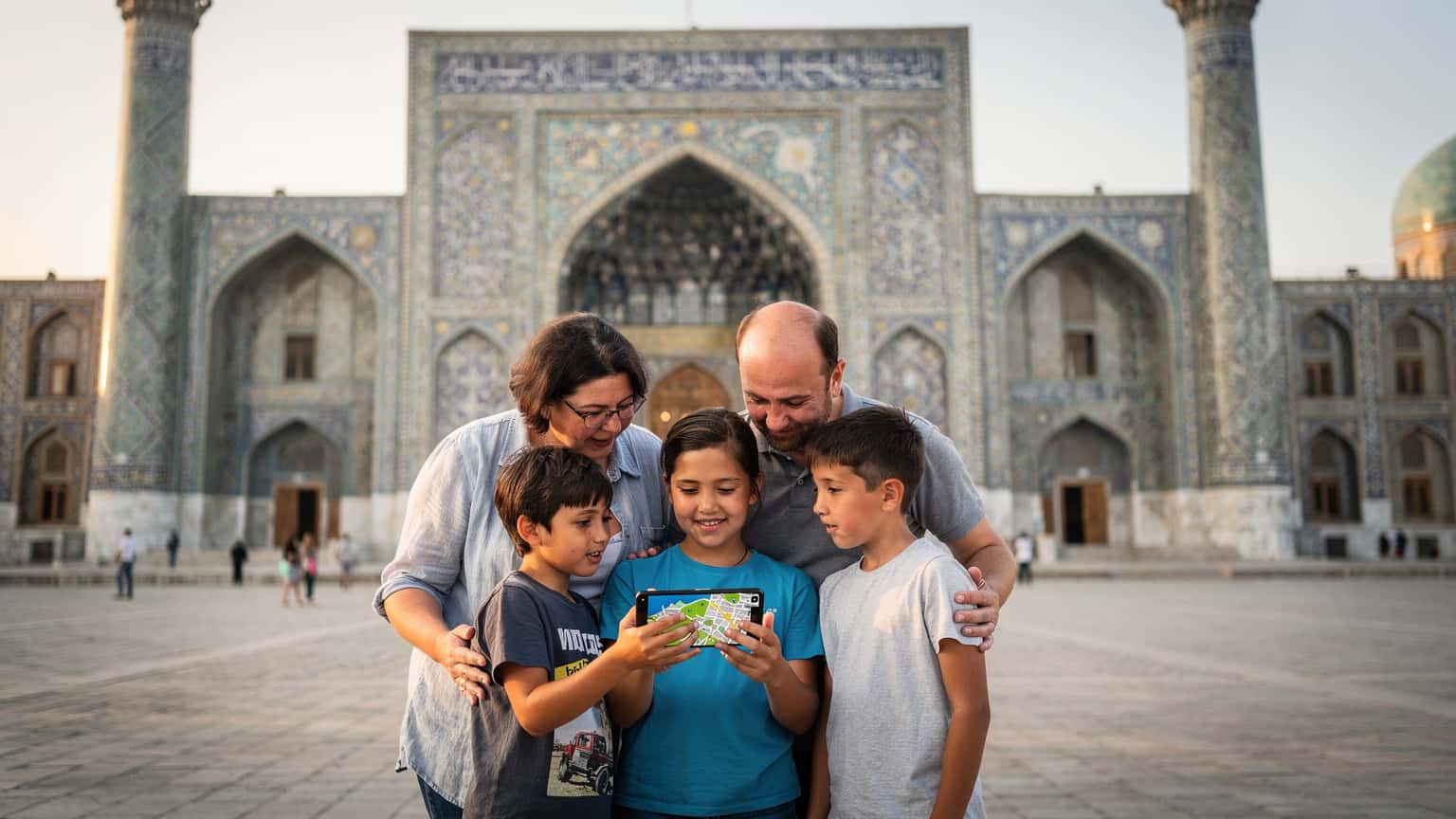 A family, including two children aged around 8 and 10, standing in front of the Registan Square in Samarkand, Uzbekistan, looking at their phones for directions, with ancient madrasahs and a clear blue sky in the background.