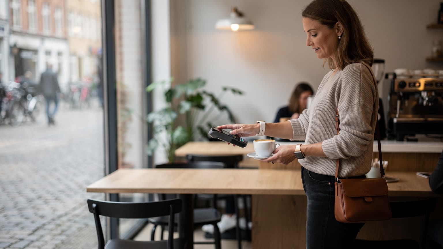En kvinde med et Apple Watch på håndleddet betaler for en kop kaffe i en hyggelig café i Københavns Indre By, hendes iPhone ligger diskret væk i tasken.
