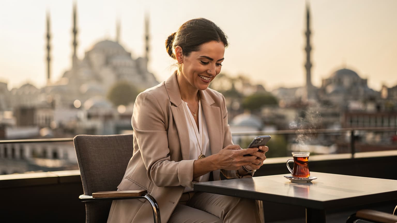 O femeie zâmbitoare navighează pe telefonul mobil pe terasa unui acoperiș din cartierul Sultanahmet, cu Hagia Sofia și Moscheea Albastră în fundal la apus, având o ceașcă de ceai turcesc alături.