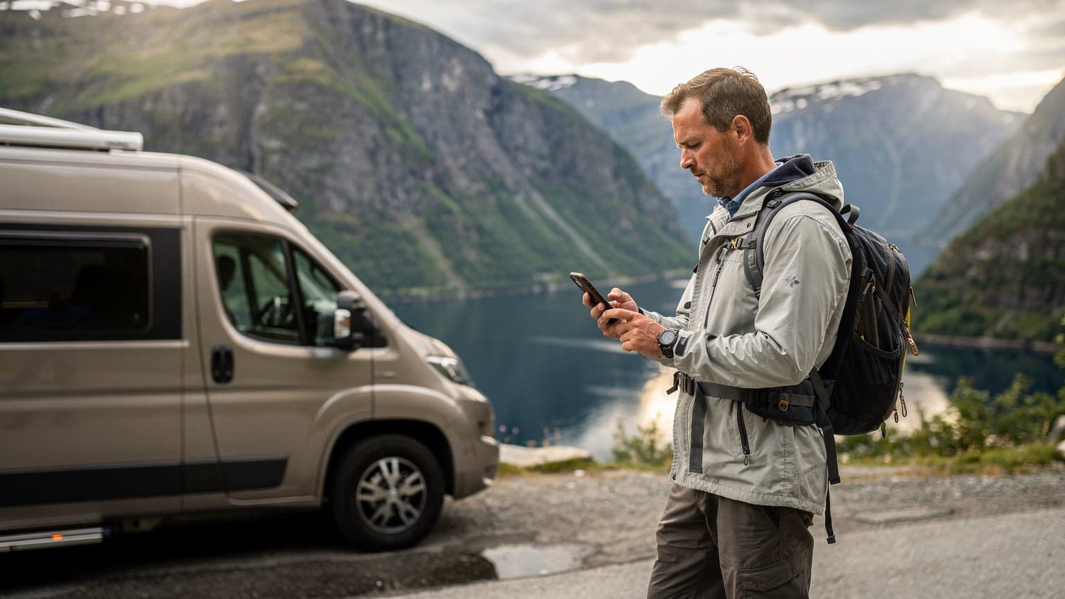Een reiziger bekijkt een digitale kaart op een smartphone, met op de achtergrond een idyllisch Noors fjordlandschap en een geparkeerde camper.