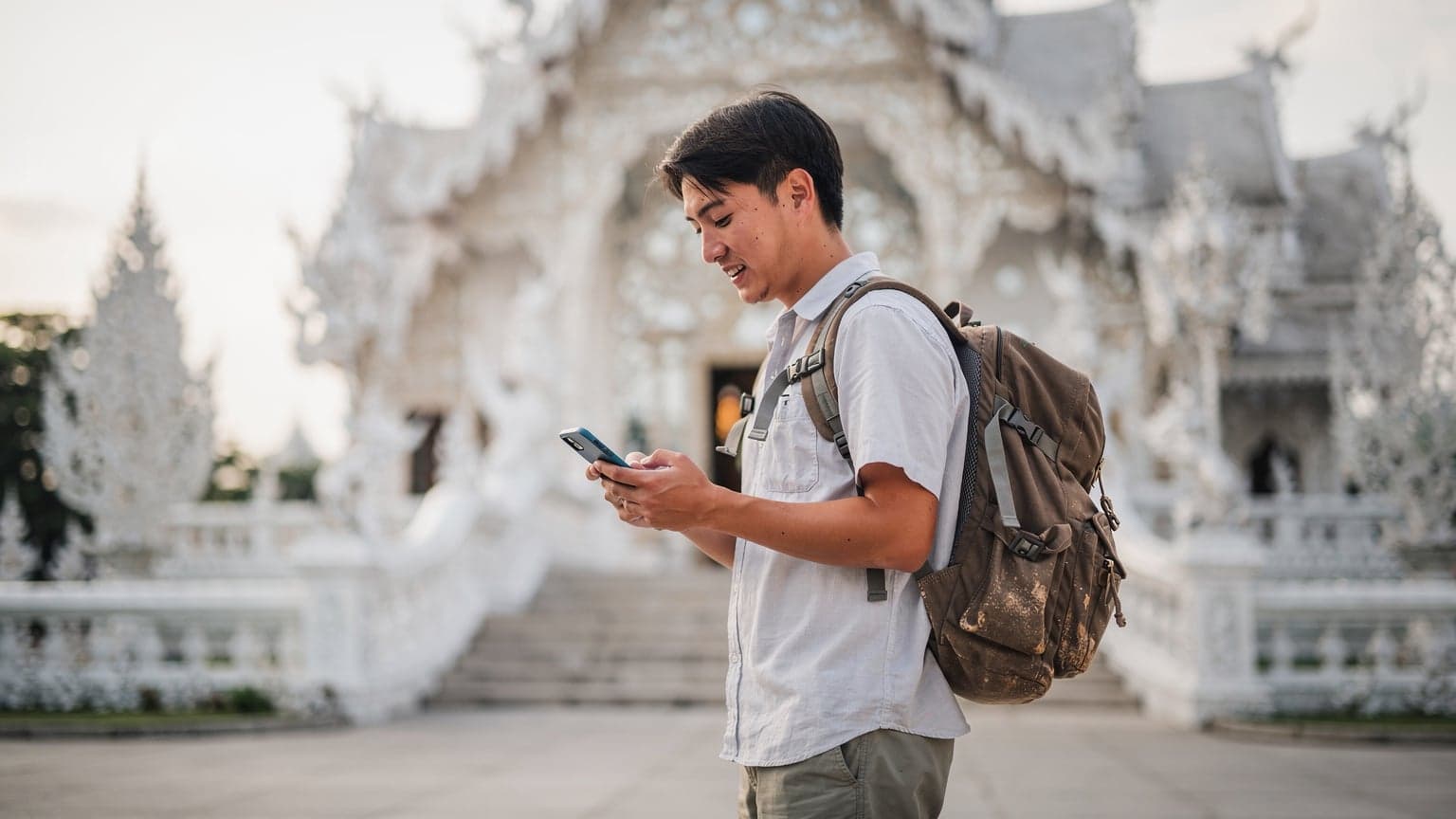 タイのチェンライのワット・ロンクン(ホワイト・テンプル)を背景に、スマートフォンで地図アプリを確認している日本人バックパッカー。