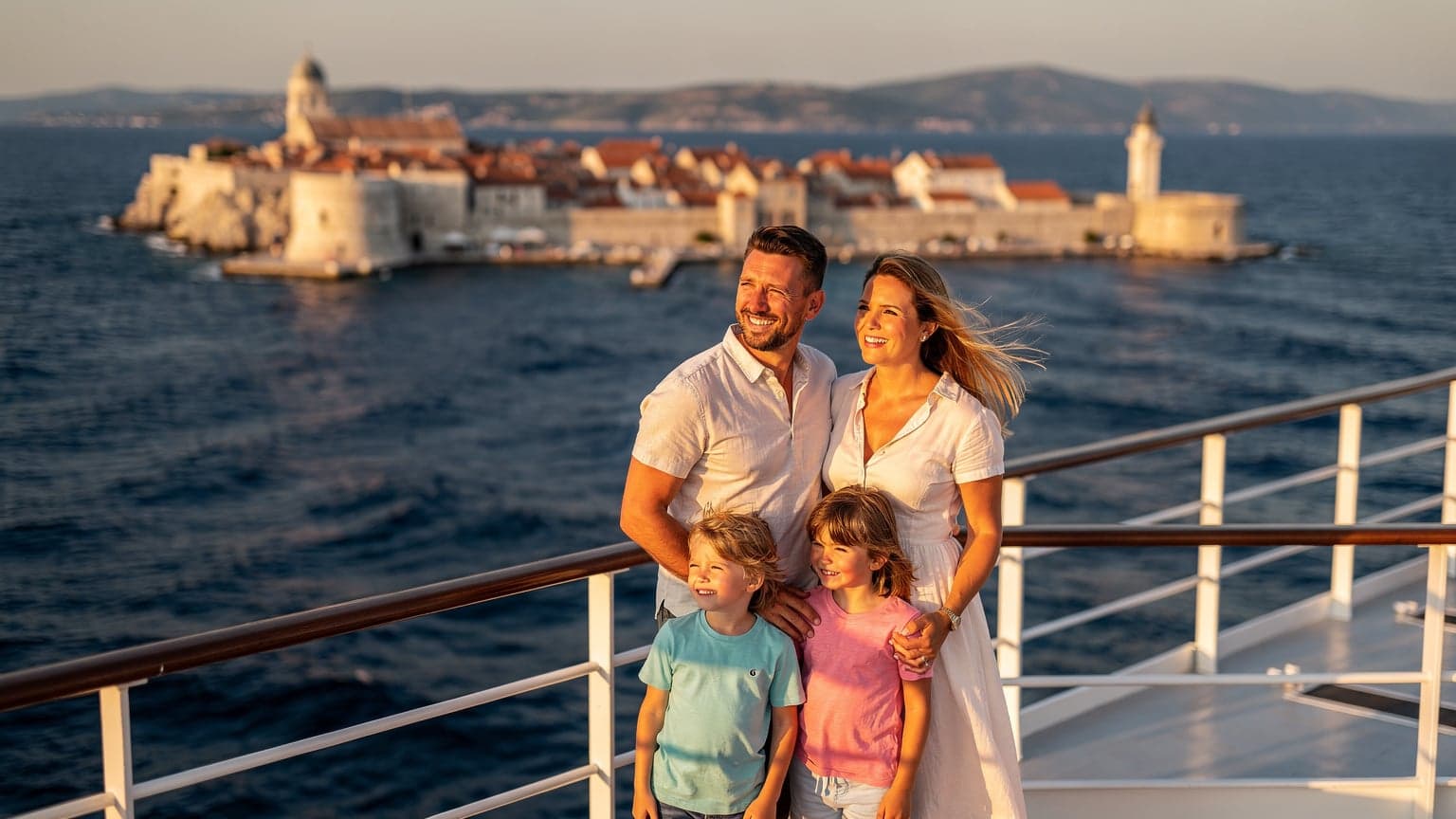 Eine Familie, bestehend aus Vater, Mutter und zwei Kindern im Alter von etwa 8 und 10 Jahren, steht lächelnd an der Reling eines Kreuzfahrtschiffes und blickt auf das tiefblaue Meer der Adria. Im Hintergrund sind die Umrisse einer malerischen Küstenstadt mit historischen Gebäuden und einem Leuchtturm zu sehen. Der Vater hält ein Smartphone in der Hand, auf dem er gerade eine Landkarte überprüft, während die Kinder gespannt auf etwas am Horizont zeigen.
