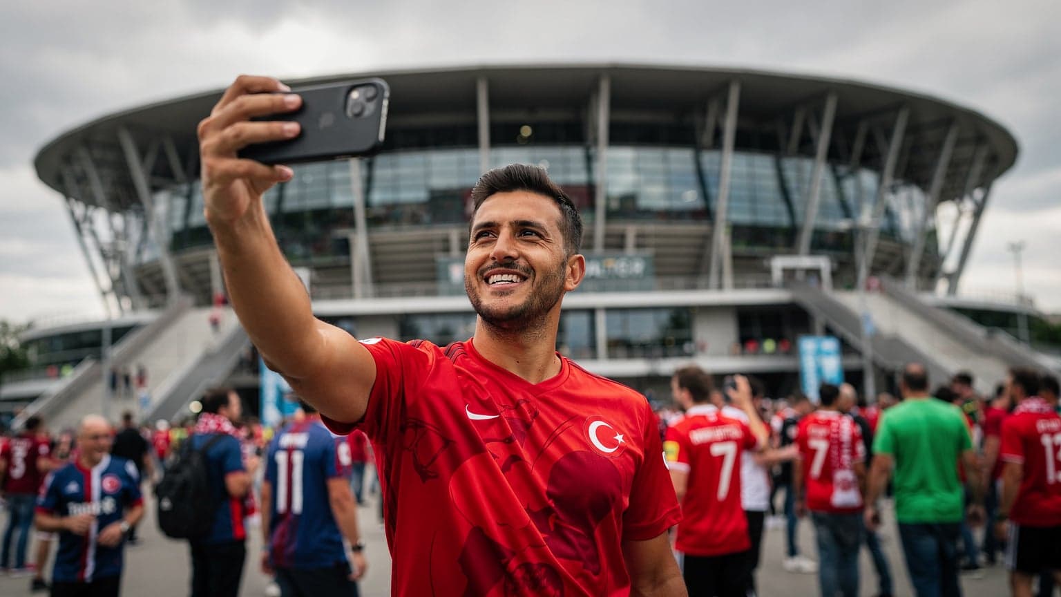 Bir Türk taraftar, Avrupa'da bir futbol stadyumunun dışında, elinde akıllı telefonuyla gülümseyerek selfi çekiyor, arkasında büyük bir stadyum ve diğer taraftarlar görünüyor.