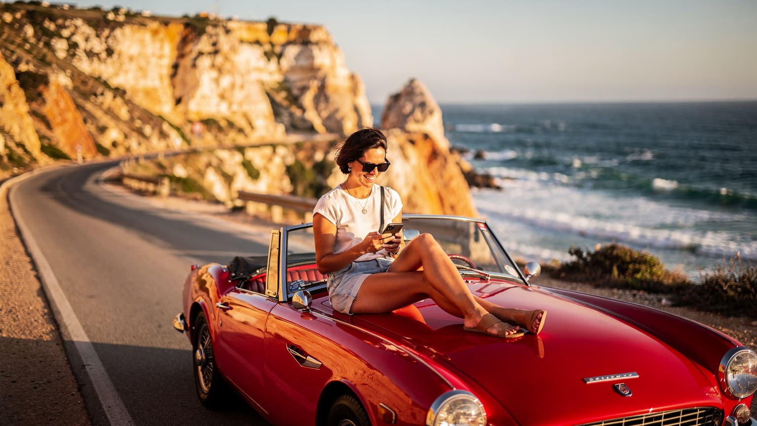 Une femme souriante, assise sur le capot d'une voiture décapotable rouge garée sur une route côtière ensoleillée du Portugal, consulte son téléphone avec une vue imprenable sur l'océan Atlantique au coucher du soleil.