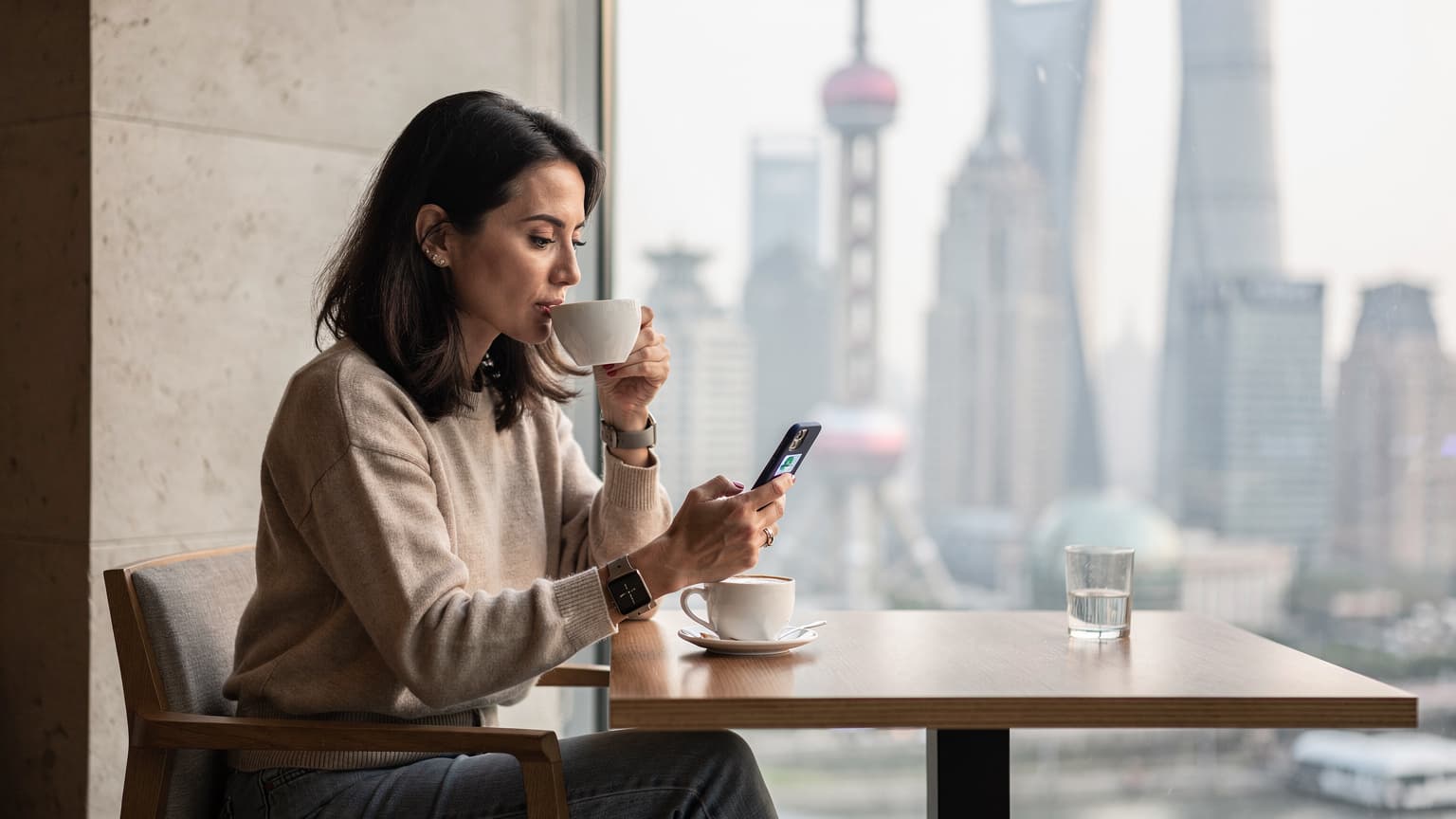 一名旅行者在上海陸家嘴金融區的咖啡館裡,手持手機查看中國移動eSIM的設置頁面,窗外是繁華的城市天際線和東方明珠電視塔,光線透過玻璃灑落在桌面上。