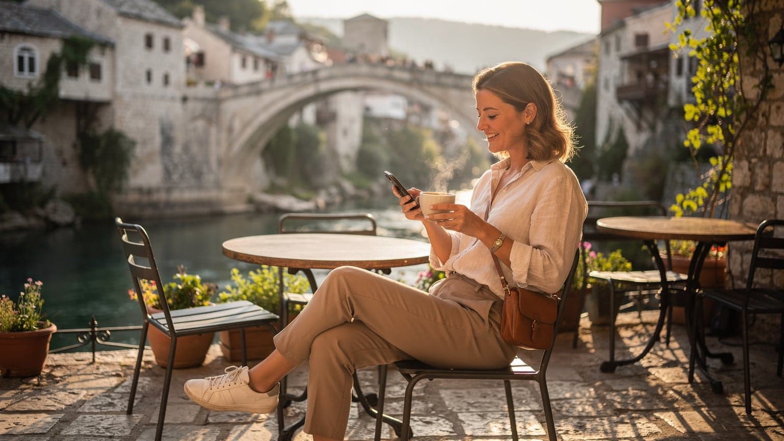 Eine Frau sitzt auf einer sonnigen Terrasse in der Altstadt von Mostar, Bosnien und Herzegowina, mit Blick auf die Neretva und die Stari Most, während sie entspannt ihr Smartphone überprüft.
