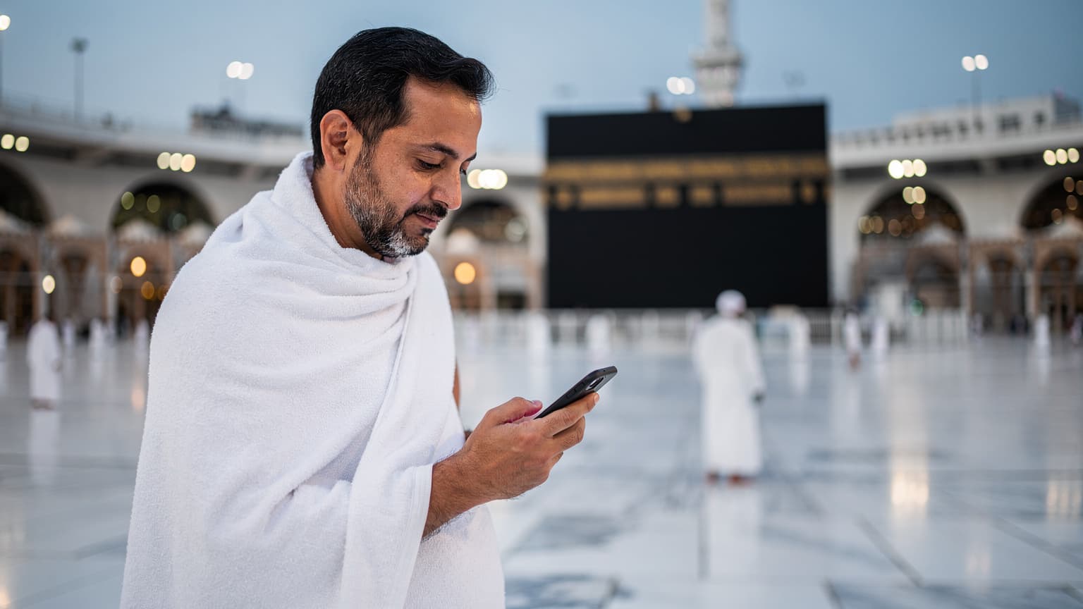 Seorang jamaah pria paruh baya mengenakan ihram sedang memegang smartphone dengan layar menyala, berdiri di depan latar belakang Masjidil Haram yang megah di Makkah, saat waktu Subuh yang tenang.