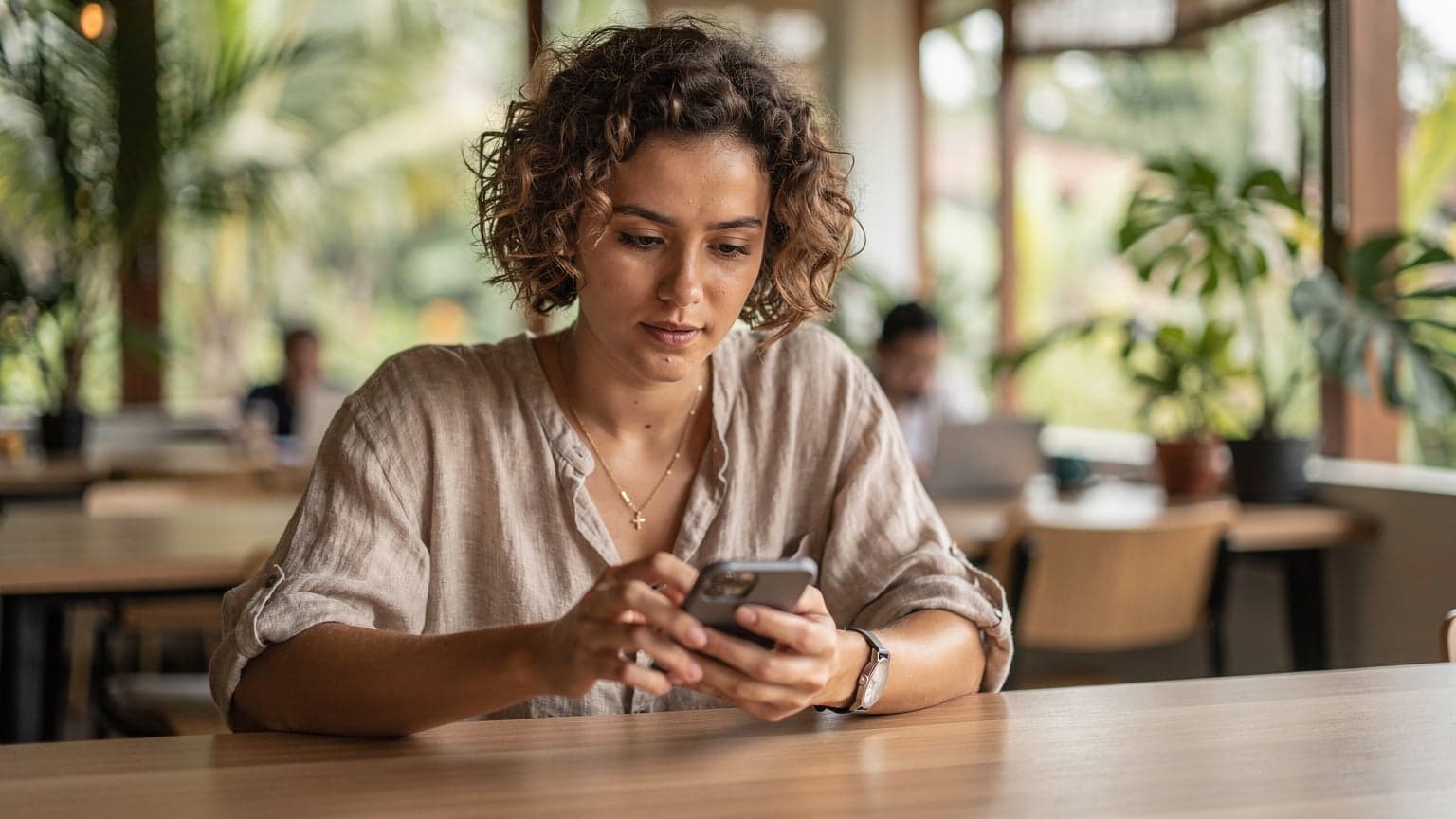 A digital nomad sits at a co-working space in Canggu, Bali, looking at her phone with a focused expression, a laptop open in front of her.