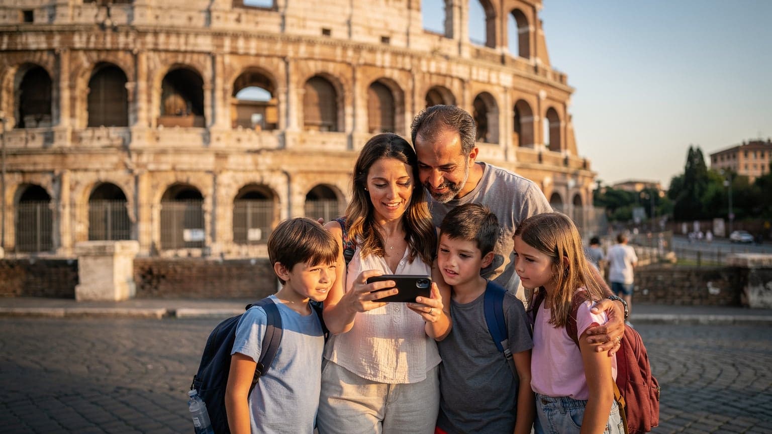 A family, including two children aged around 8 and 10, standing in front of the Colosseum in Rome, looking at a smartphone held by the mother, possibly checking directions or a digital guide.