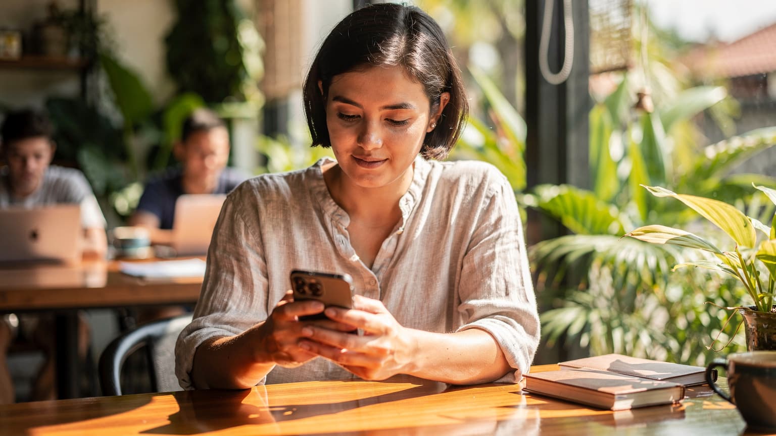 A digital nomad sits at a wooden table in a busy co-working cafe in Canggu, Bali, looking intently at her iPhone screen while activating an eSIM, with a tropical plant visible in the background.