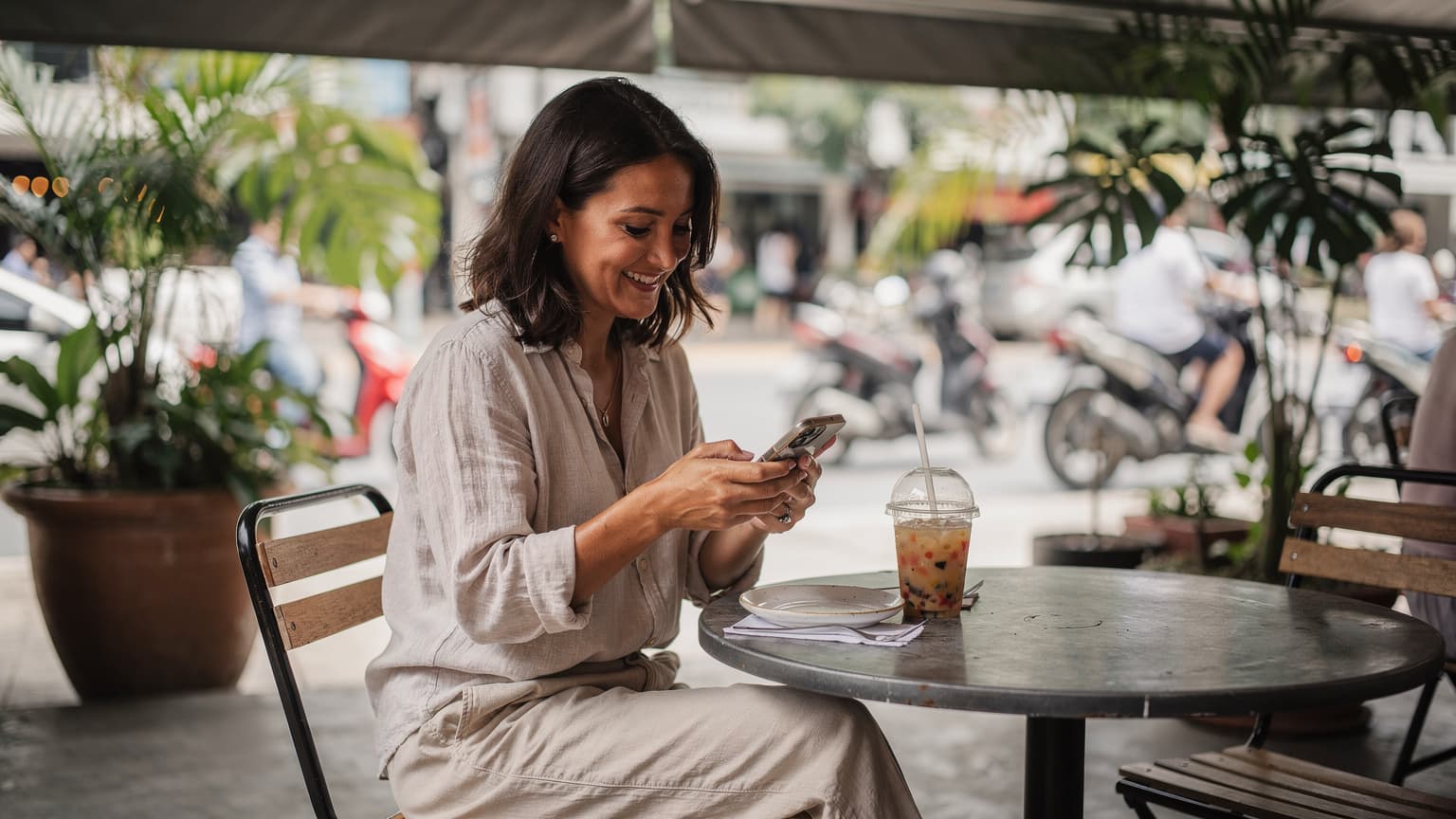 Une femme souriante, assise à la terrasse d'un café animé à Bangkok, consulte son téléphone portable avec une boisson fraîche à portée de main, le tout sous une lumière douce du matin.