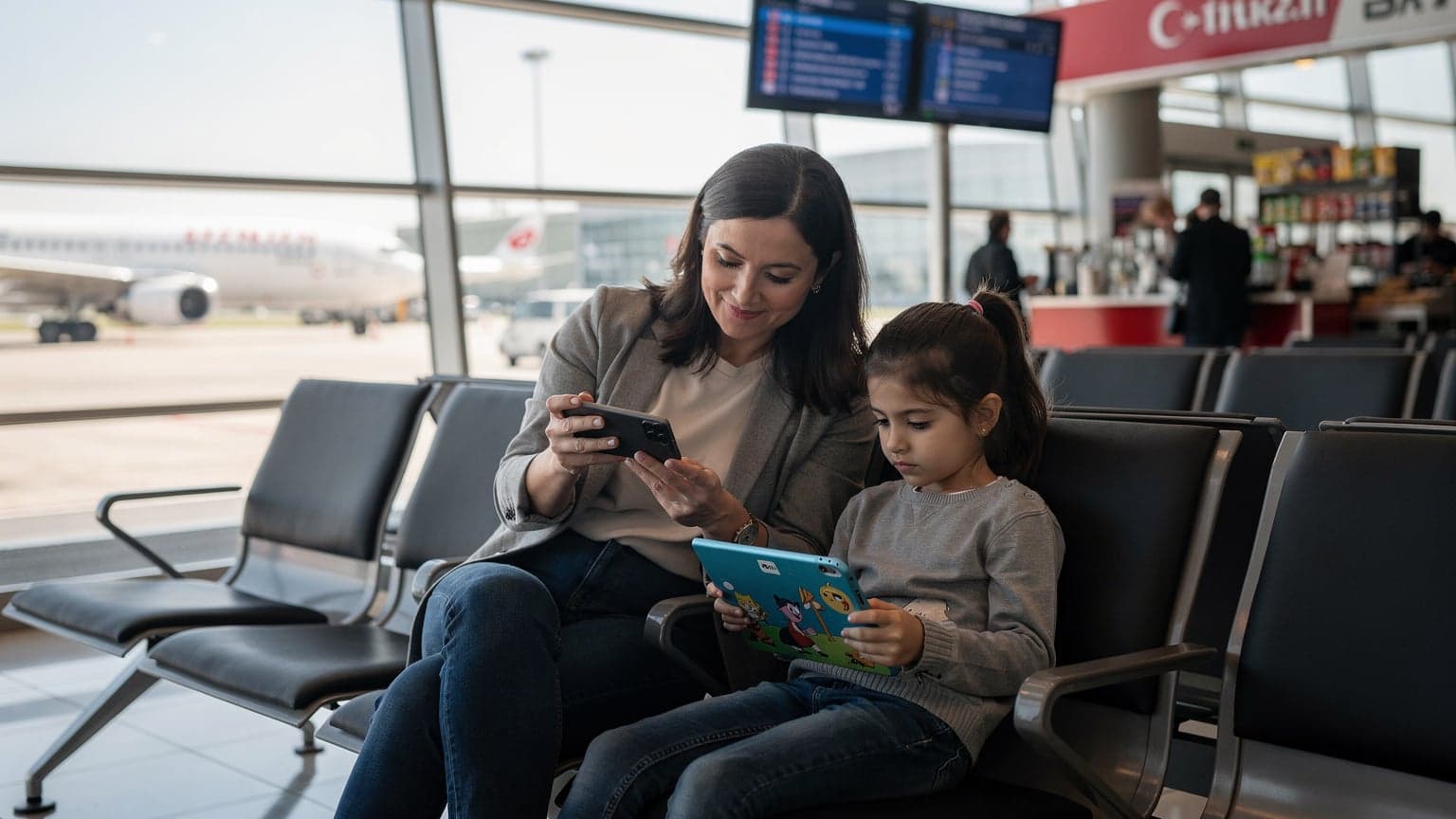 İstanbul Havalimanı'nda, uçağa binmeden önce tabletinde çizgi film izleyen bir çocuk ve yanında telefonundan eSIM paketi kontrol eden bir anne, bavulların arasında oturuyor.