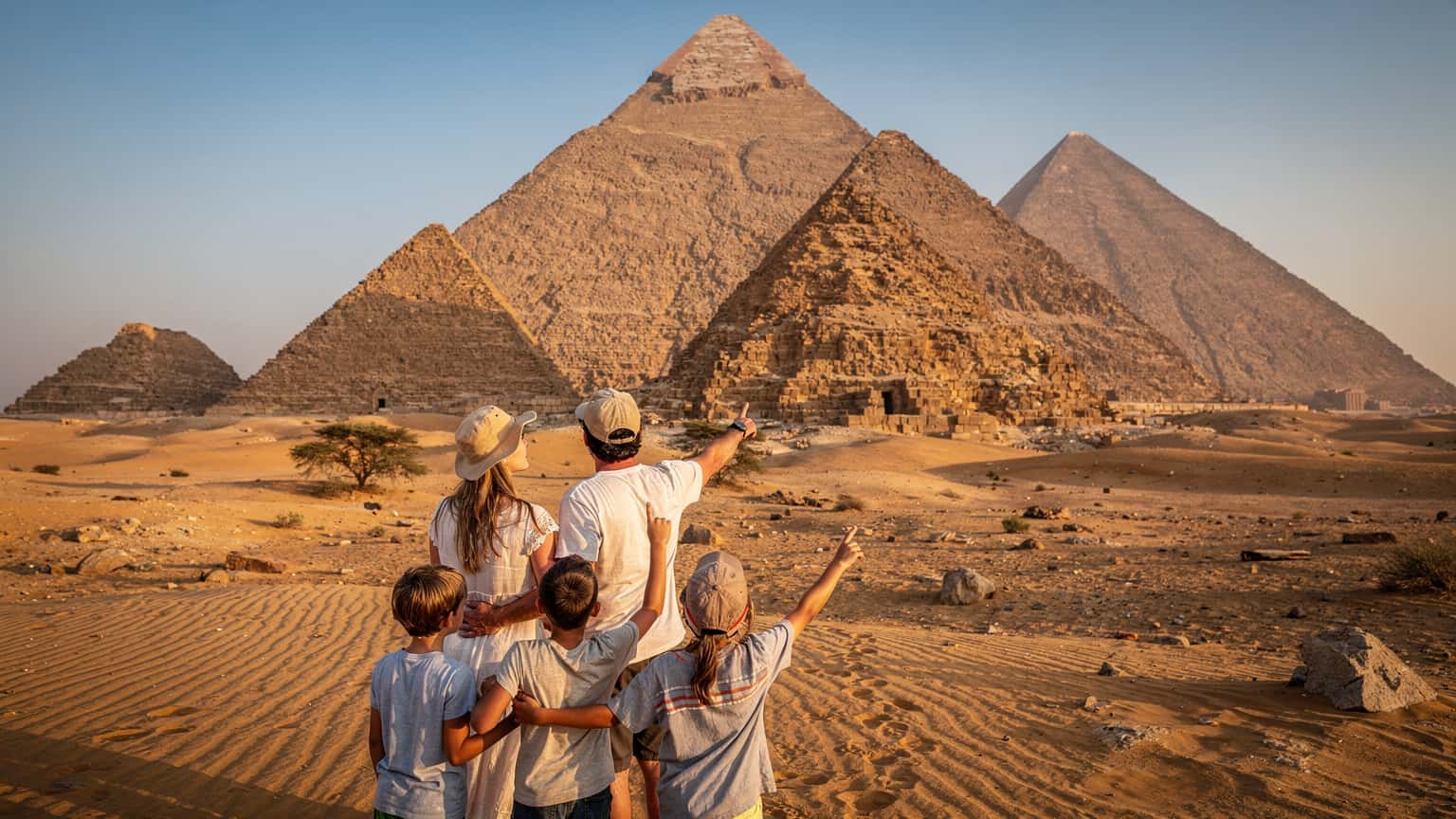 A family with two children, aged around 8 and 10, standing in awe before the ancient pyramids of Meroë in Sudan under a clear blue sky, a sense of wonder and history in their expressions.