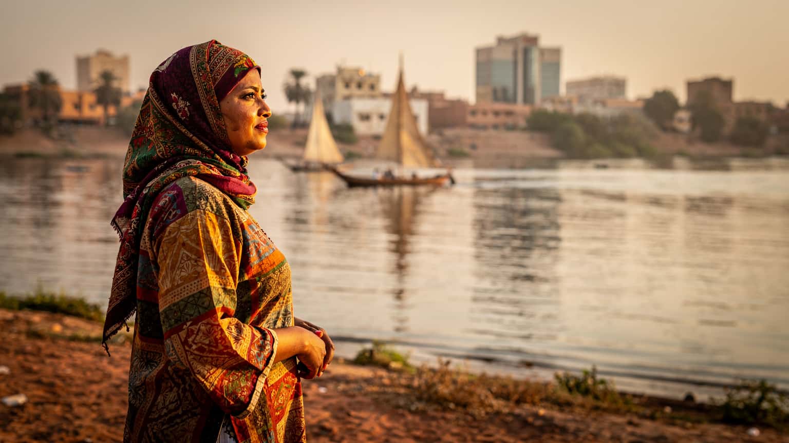 A woman wearing a vibrant headscarf gazes out over the Nile River in Khartoum, Sudan, at sunset, with traditional feluccas silhouetted against the glowing sky.