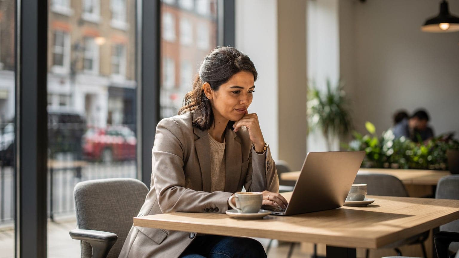 Een vrouw met een laptop op schoot, nippend aan een koffie in een lichtovergoten coworking space in Shoreditch, Londen, met de skyline op de achtergrond.
