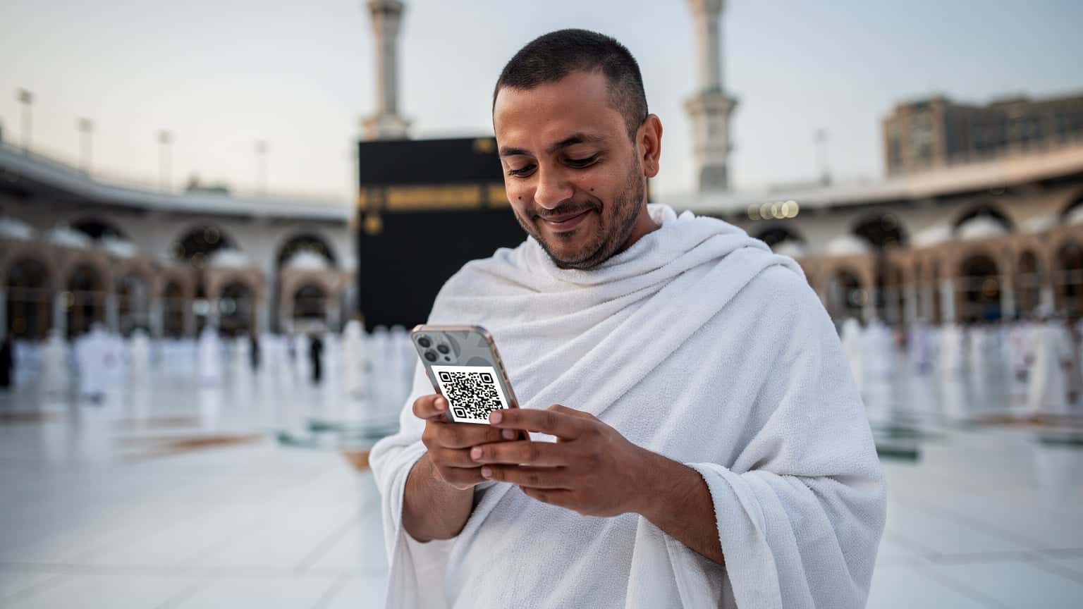 A pilgrim in traditional Ihram attire holding a smartphone with a data plan QR code on the screen, standing near the Grand Mosque in Mecca, Saudi Arabia.