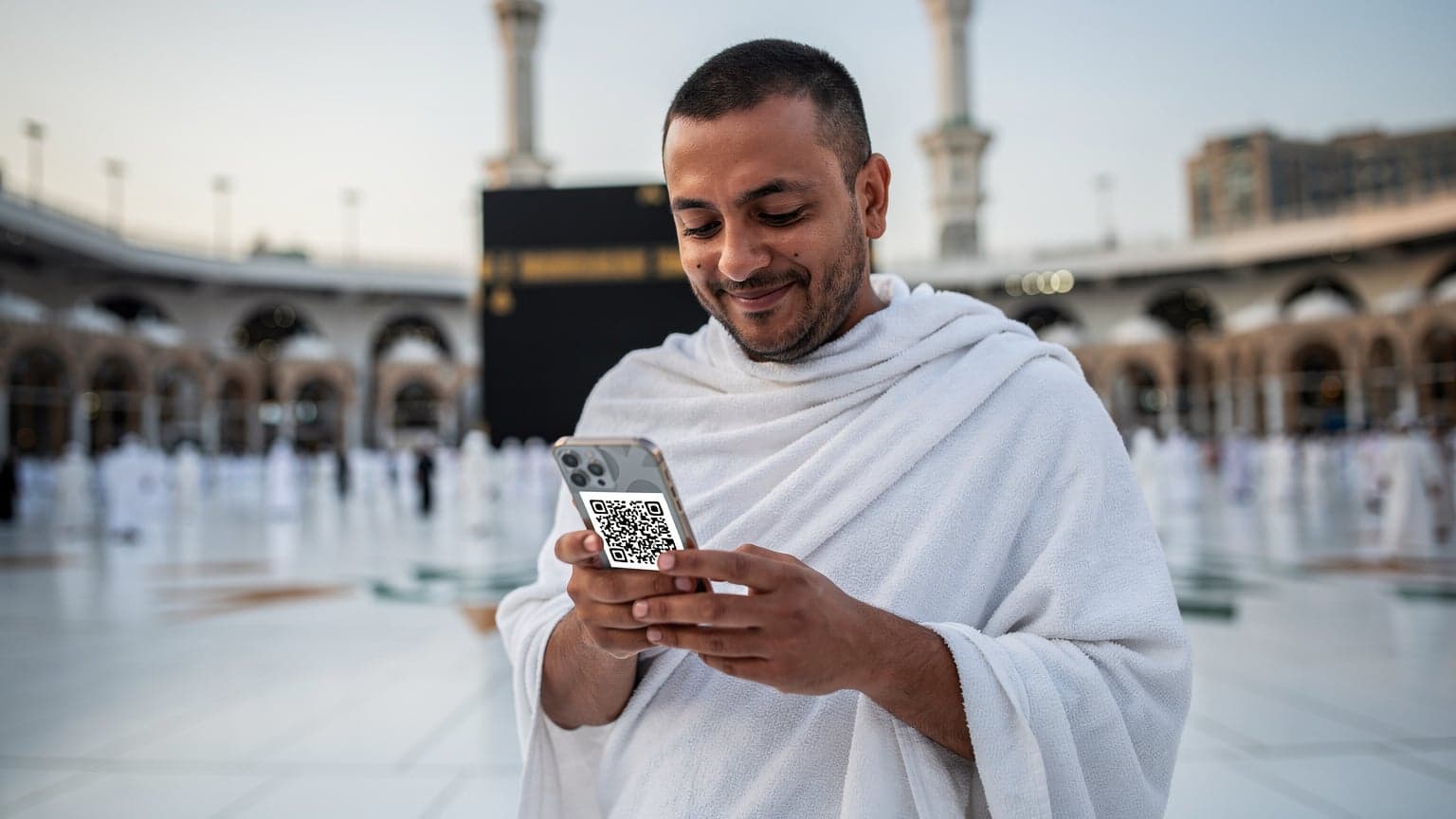 A pilgrim in traditional Ihram attire holding a smartphone with a data plan QR code on the screen, standing near the Grand Mosque in Mecca, Saudi Arabia.