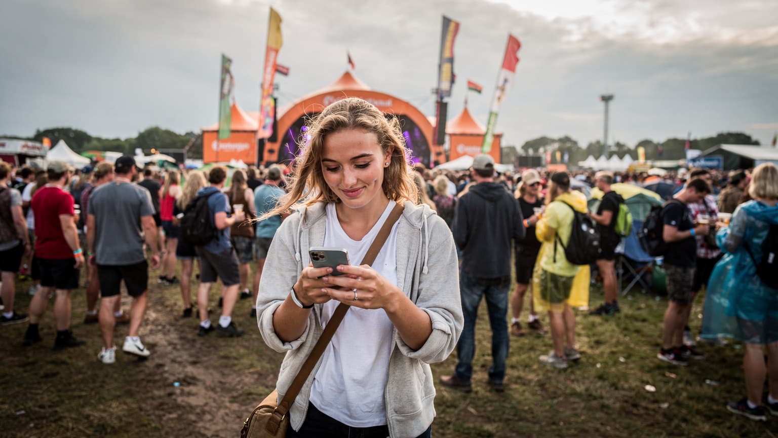 En ung kvinde navigerer med sin smartphone i en stor menneskemængde på Roskilde Festivalens orange scene, med farverige flag og telte i baggrunden.
