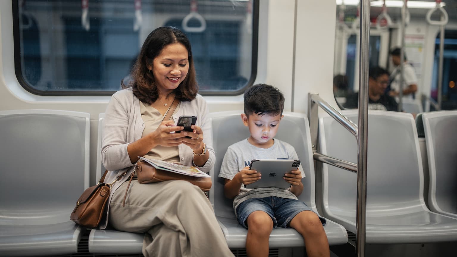 Seorang ibu tersenyum sambil memegang telefon pintar dan seorang anak lelaki melihat tablet di dalam MRT Singapura yang bersih dan moden.