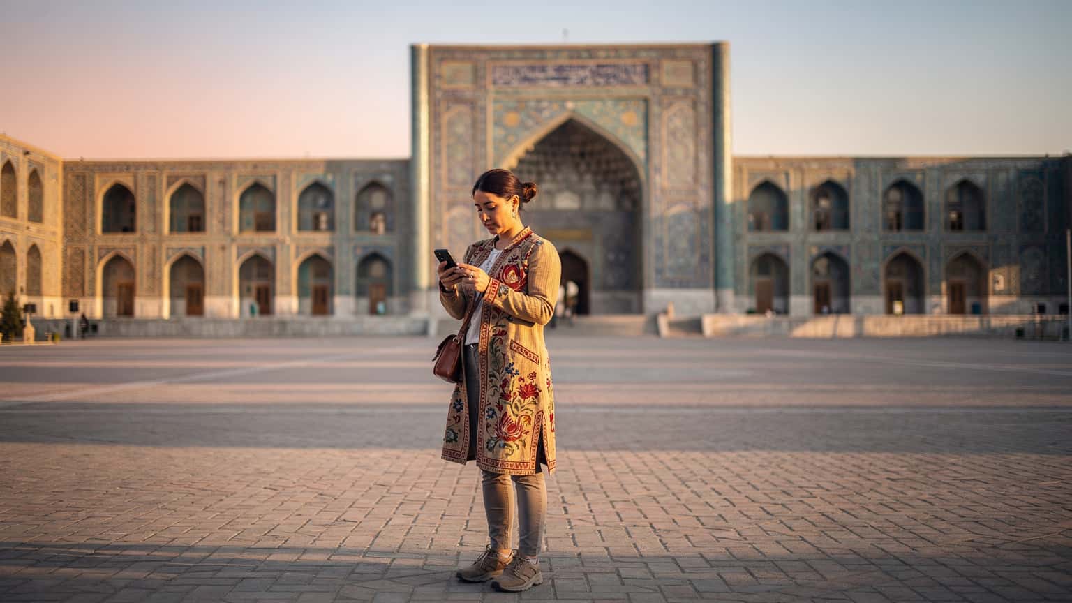A traveler in a traditional Uzbek suzani coat, holding a smartphone with a vibrant map displayed, stands in front of the Registan Square in Samarkand at dawn.