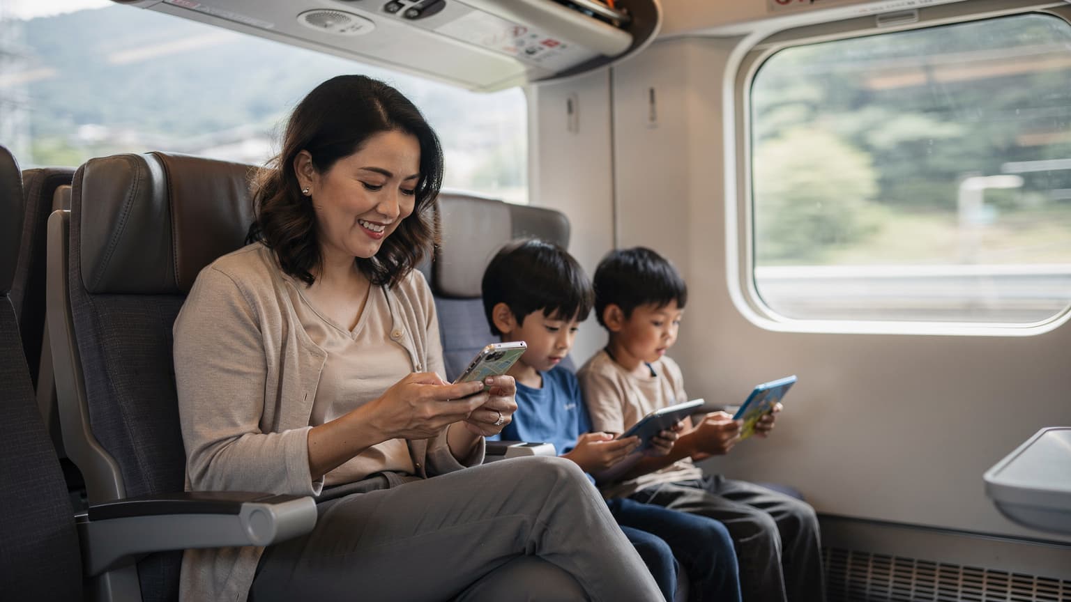 Seorang ibu tersenyum sambil melihat peta digital di telefonnya, dikelilingi oleh dua orang anak yang sedang bermain dengan tablet di tangan, di dalam sebuah kereta api laju Shinkansen yang moden di Jepun.