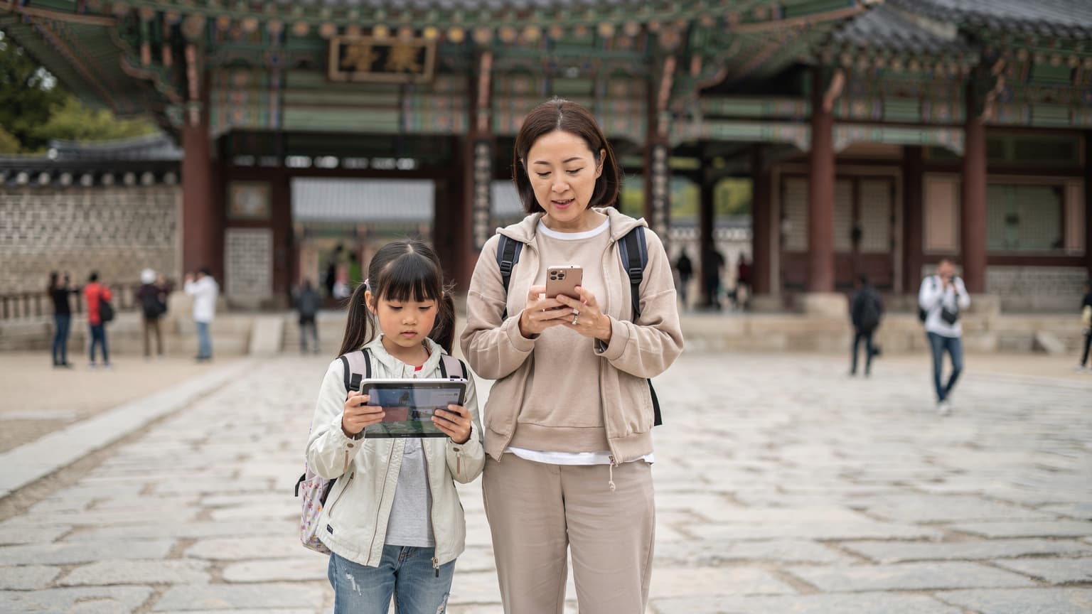 ソウルの景福宮の敷地内で、母親がスマートフォンで地図を確認し、隣で幼い娘がタブレットを操作している様子。