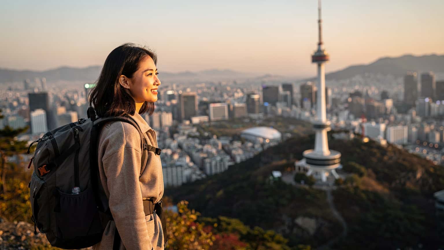 O femeie zâmbitoare, cu un rucsac ușor, privește spre orizontul orașului Seul, cu turnul Namsan în fundal, la apus.