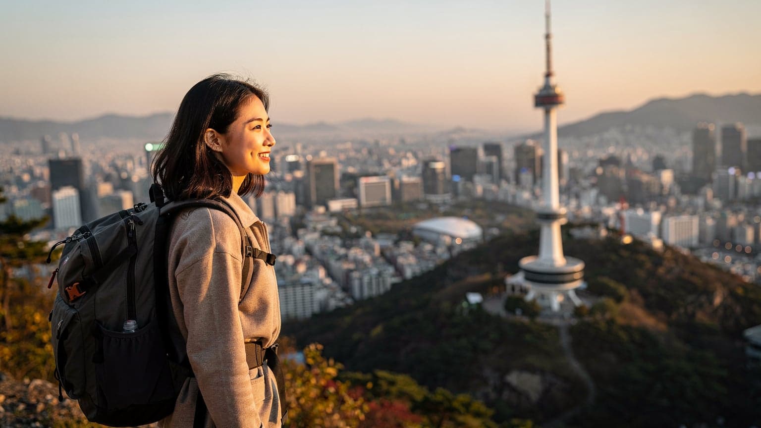 O femeie zâmbitoare, cu un rucsac ușor, privește spre orizontul orașului Seul, cu turnul Namsan în fundal, la apus.