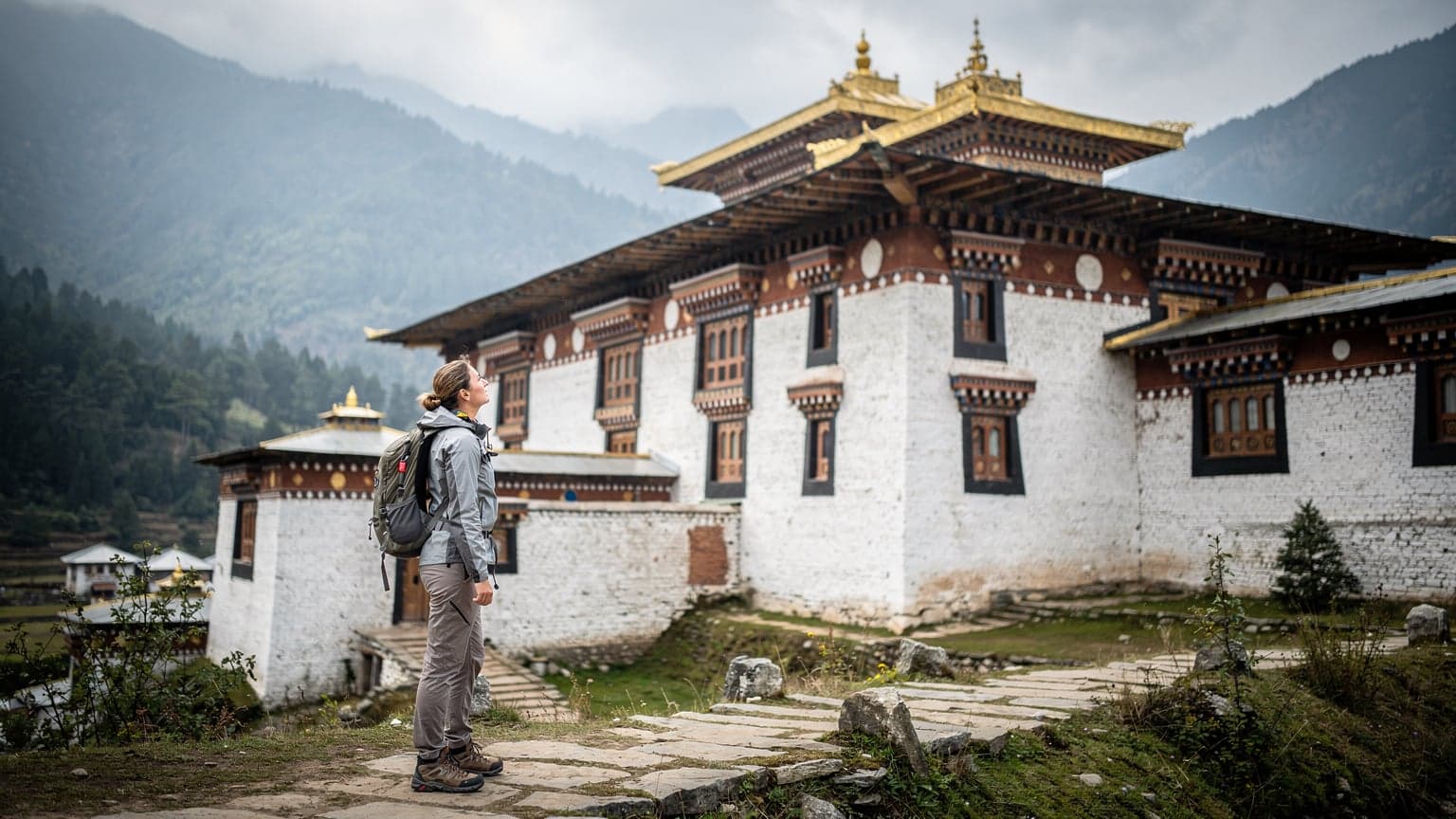 A solo female traveler observing the intricate architecture of a traditional Bhutanese monastery, with prayer flags fluttering gently in the crisp mountain air under a soft morning sky.