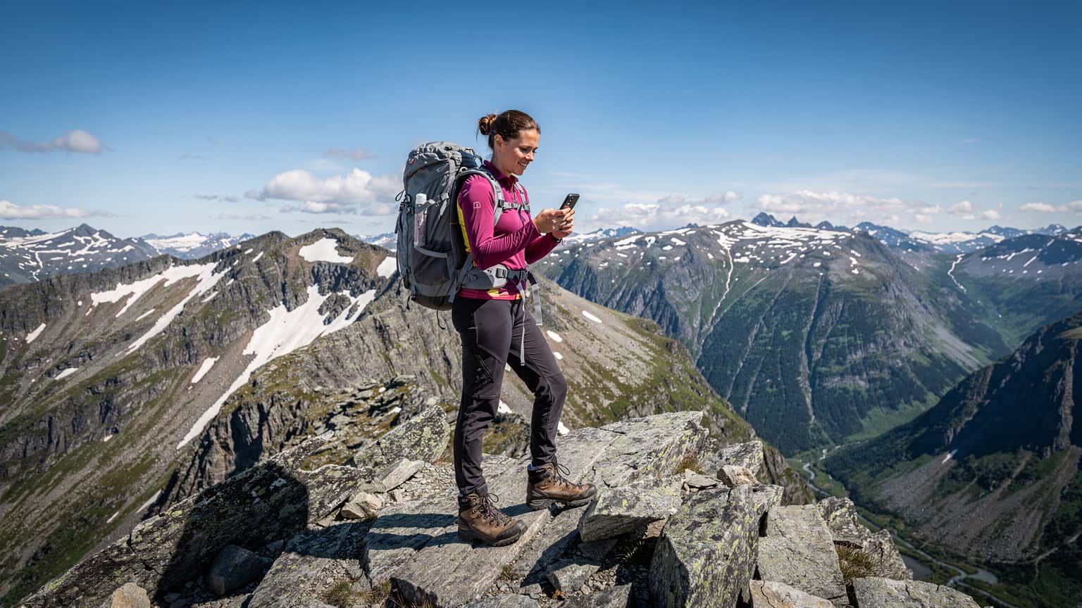 En kvinne med ryggsekk og hikestøvler står på en fjelltopp i Jotunheimen, med en mobiltelefon i hånden som viser et kart, omgitt av majestetiske fjelltopper og en klar, blå himmel.