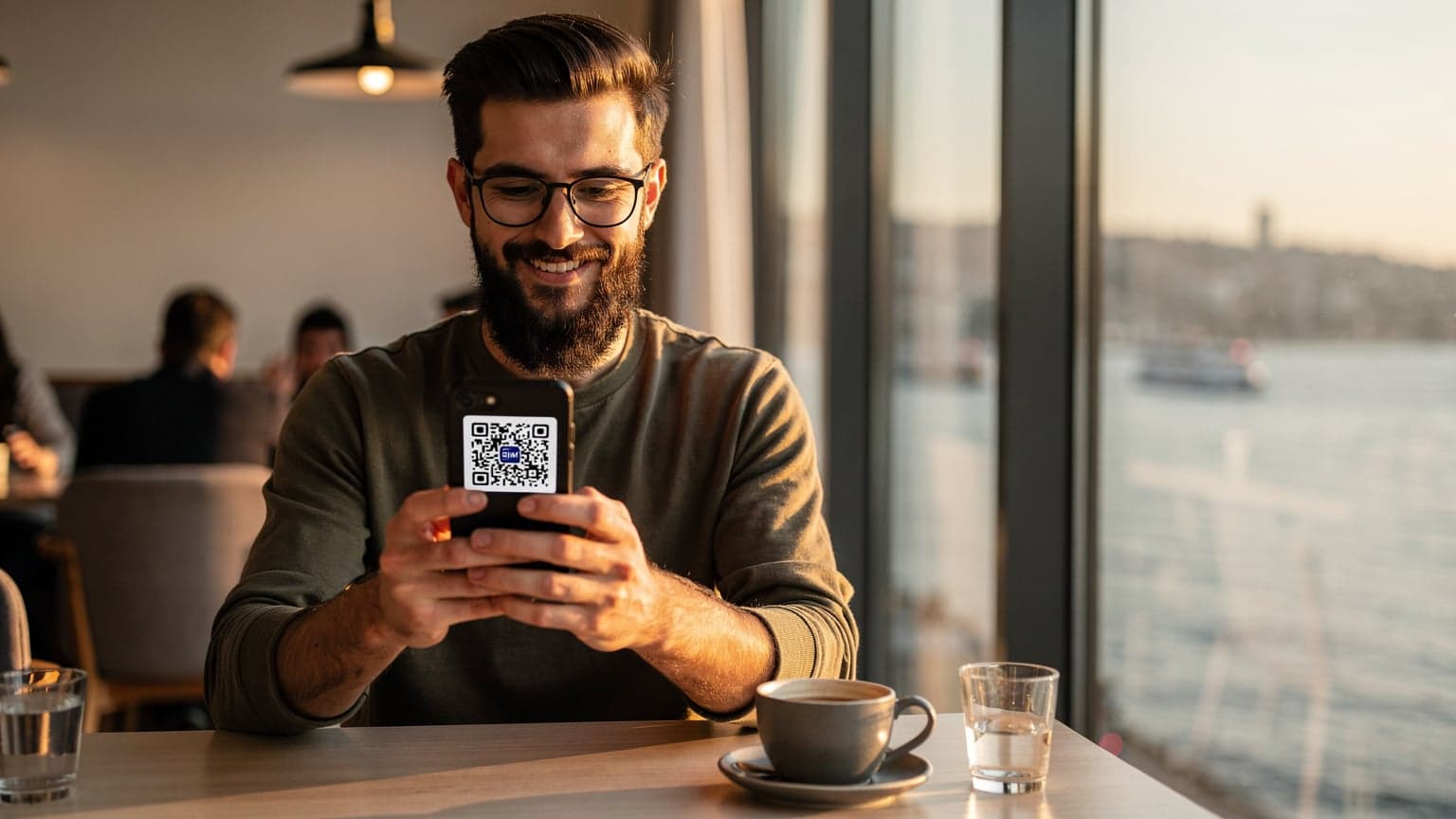 İstanbul Boğazı'na bakan modern bir kafede oturan genç bir adam, dizüstü bilgisayarının yanında telefonunda eSIM aktivasyonu yapıyor.