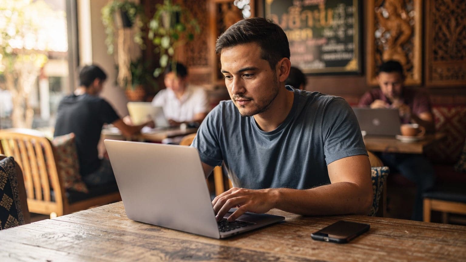 Ein digitaler Nomade sitzt in einem geschäftigen Café in Chiang Mai, Thailand, mit einem Laptop und einem Smartphone, während er eine Tasse Kaffee trinkt und konzentriert arbeitet.