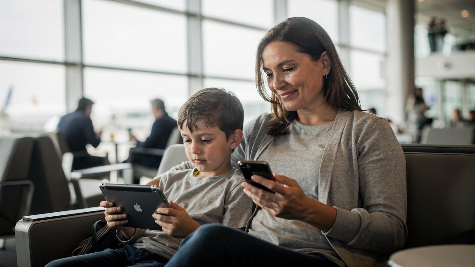 Une mère souriante et son enfant regardent un iPad sur les genoux de l'enfant dans un salon d'aéroport moderne, avec des bagages à main à proximité, la mère tenant un iPhone à la main.