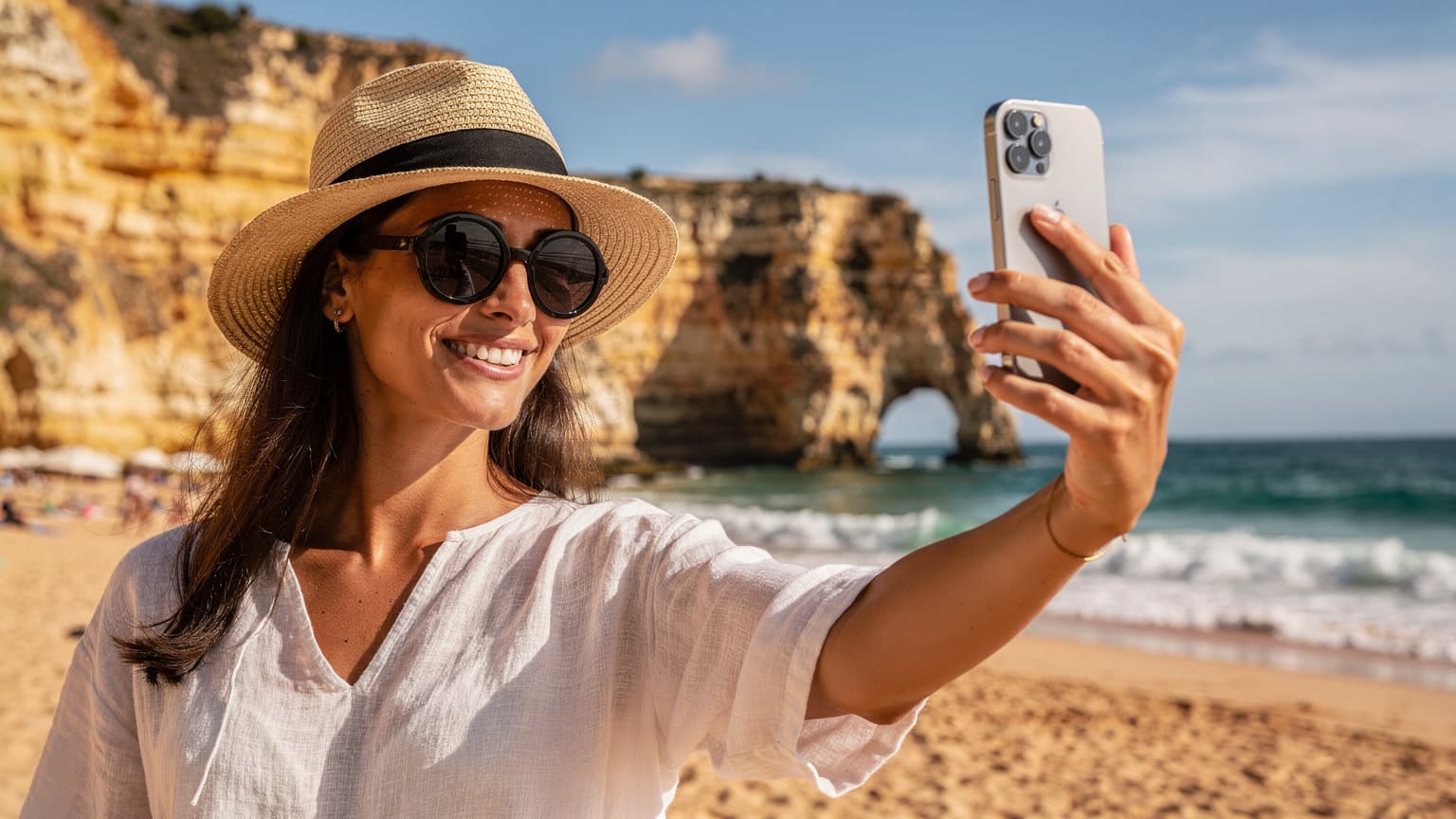Uma mulher sorridente, com cabelos escuros, usando um chapéu de palha e óculos de sol, segura um iPhone 18 na mão enquanto tira uma selfie com o mar azul-turquesa e falésias douradas do Algarve ao fundo, sob o sol forte da tarde em Portugal.