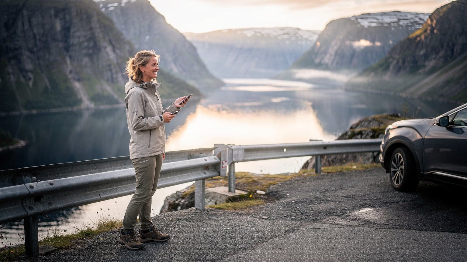 Een vrouw met zonnebril en een smartphone in de hand kijkt naar een uitgestrekt fjordlandschap in Noorwegen, haar auto geparkeerd aan de kant van een kronkelende weg, met de zon die net boven de bergen uitkomt.