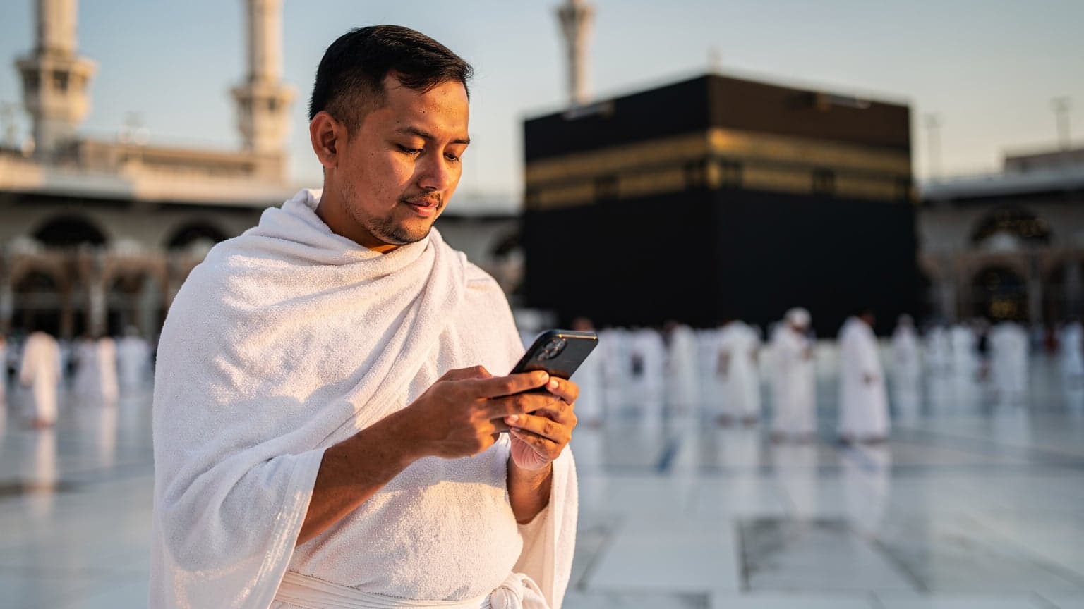 Seorang jemaah haji Malaysia sedang memeriksa telefon bimbitnya yang dilengkapi eSIM di luar Masjidil Haram di Makkah, dengan latar belakang Kaabah di kejauhan, pada waktu subuh yang tenang.