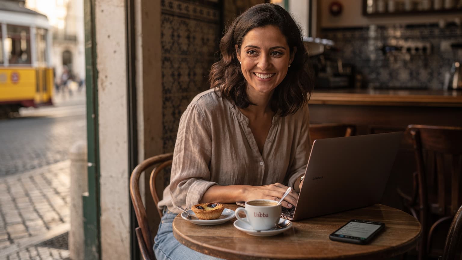 Uma mulher sorridente, com cerca de 30 anos, sentada em um café movimentado no bairro da Baixa, em Lisboa, a trabalhar no seu laptop com o telemóvel ao lado, enquanto desfruta de um pastel de nata e um café. A luz da manhã entra pelas janelas e ilumina a cena.