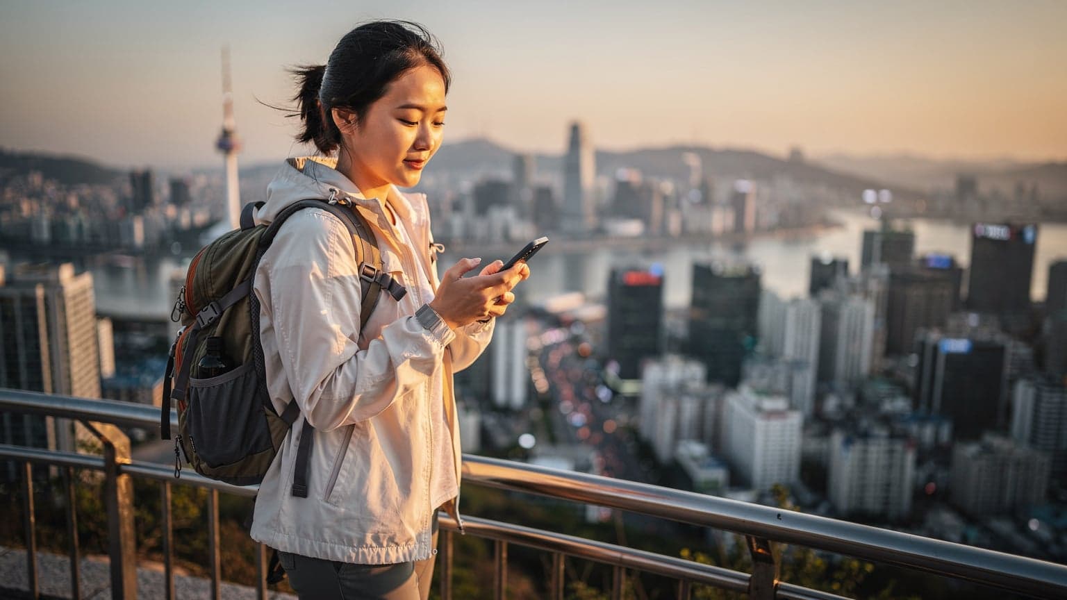 Eine junge Frau mit Rucksack blickt auf die beleuchtete Skyline von Seoul bei Sonnenuntergang, während sie ihr Smartphone überprüft.