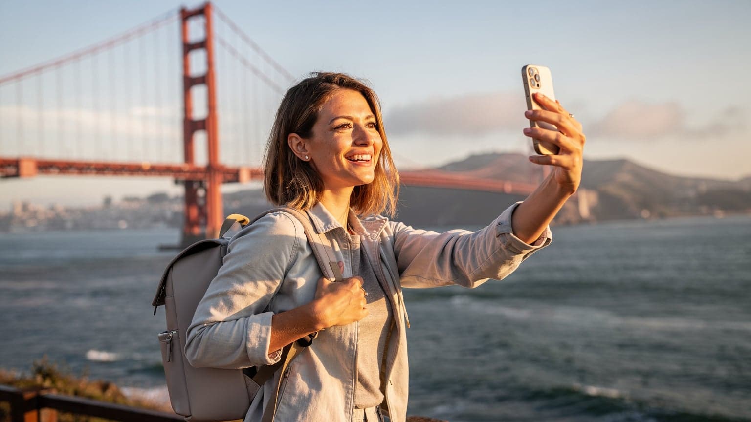 Une voyageuse souriante prend un selfie devant le pont du Golden Gate à San Francisco au lever du soleil, sa main tenant son téléphone affichant un fond d'écran clair.