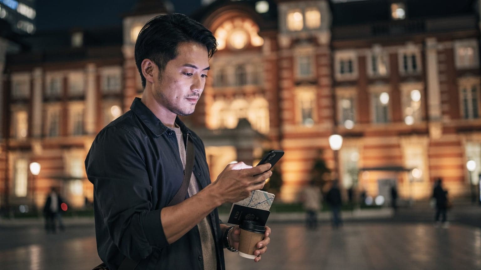 東京駅丸の内口の夜景を背景に、スマートフォンで地図アプリを見ている30代の日本人男性旅行者。手元にはパスポートと、もう片方の手には小さなコーヒーカップを持っている。