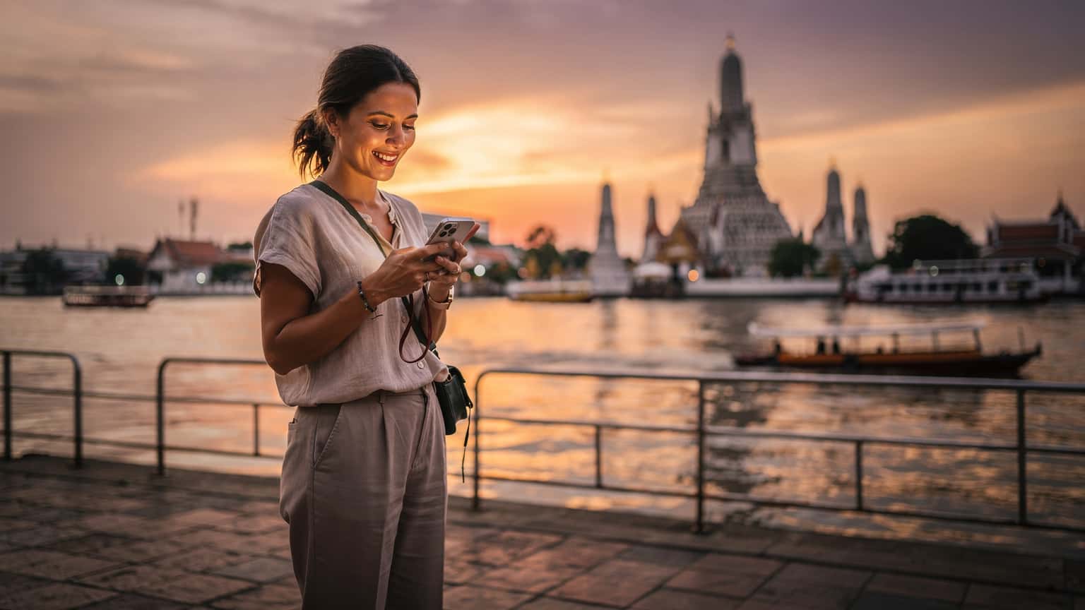 Une voyageuse souriante consulte son téléphone avec une eSIM active devant le Wat Arun au coucher du soleil à Bangkok, Thaïlande.