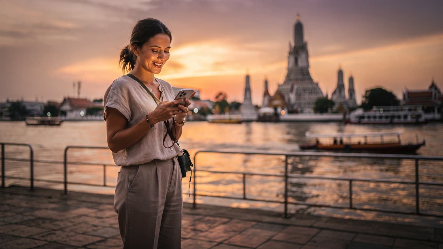 Une voyageuse souriante consulte son téléphone avec une eSIM active devant le Wat Arun au coucher du soleil à Bangkok, Thaïlande.