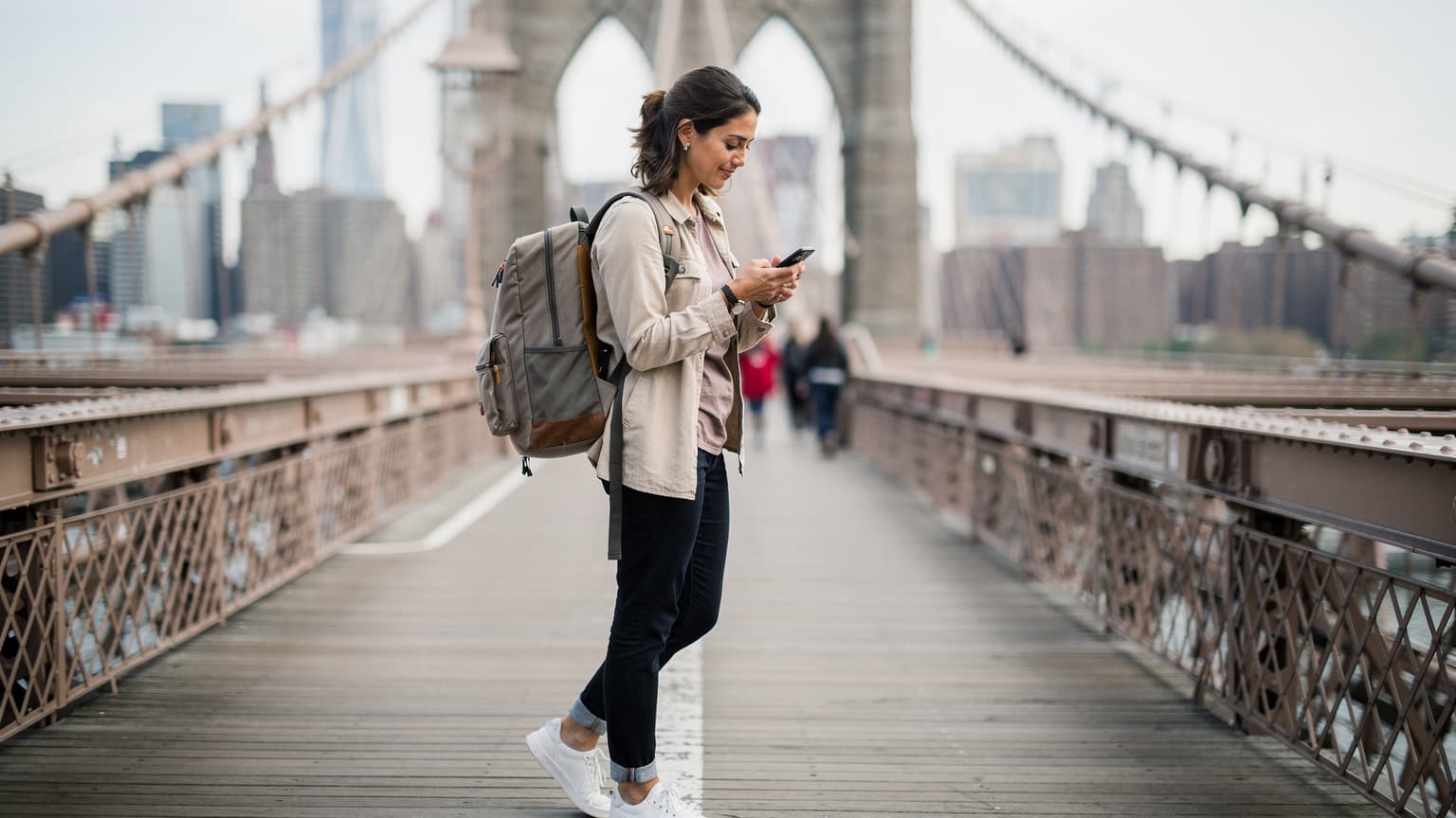 Una donna italiana con uno smartphone in mano consulta una mappa digitale mentre si trova sul ponte di Brooklyn a New York City, con lo skyline di Manhattan sullo sfondo.