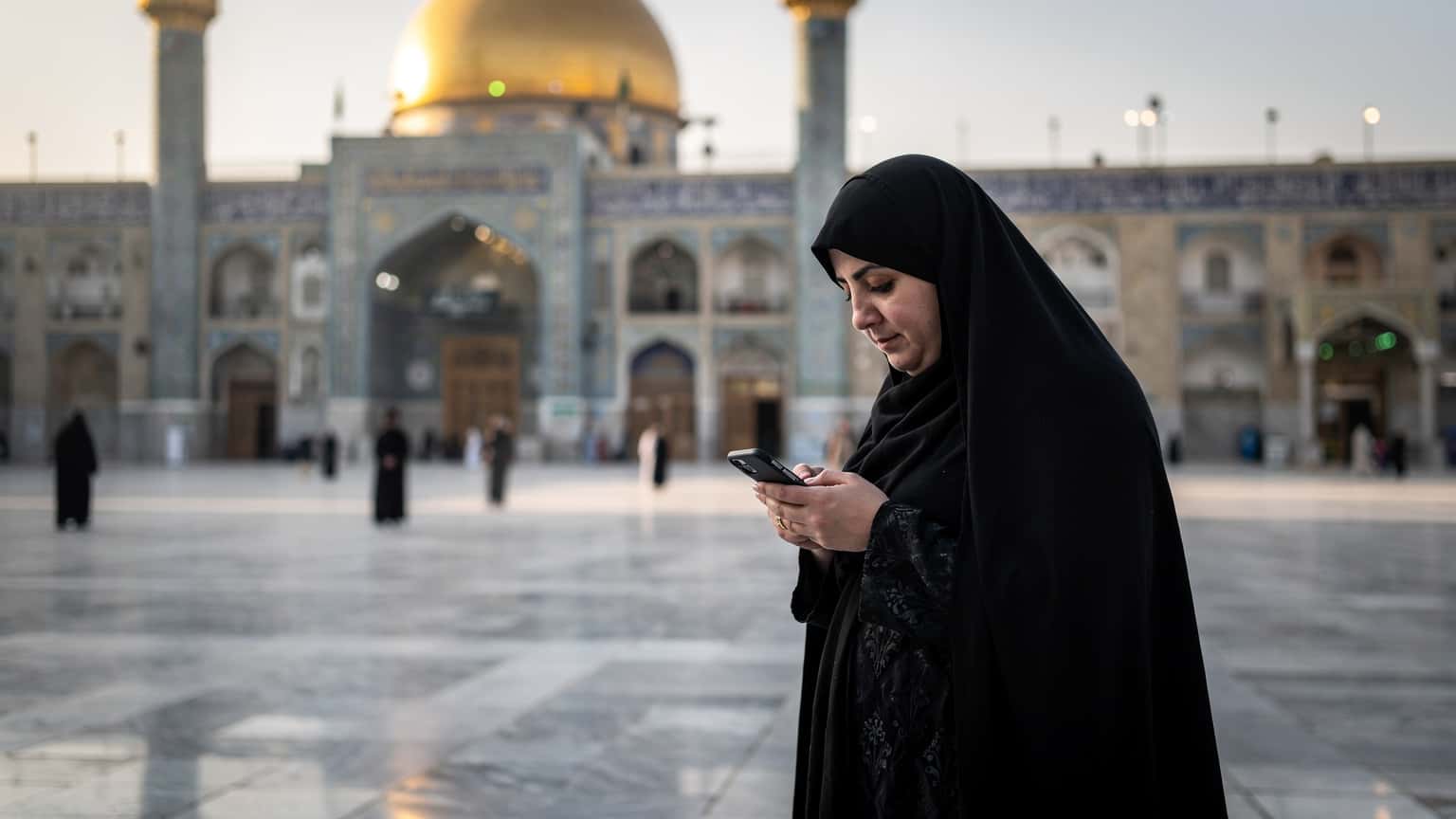زنی با حجاب در حال استفاده از تلفن همراه خود در صحن حرم مطهر امام علی در نجف، با معماری اسلامی زیبا در پس زمینه و نور ملایم صبحگاهی.