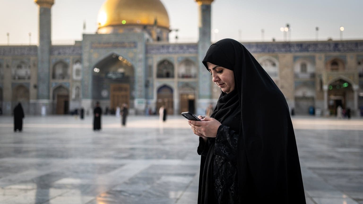 زنی با حجاب در حال استفاده از تلفن همراه خود در صحن حرم مطهر امام علی در نجف، با معماری اسلامی زیبا در پس زمینه و نور ملایم صبحگاهی.