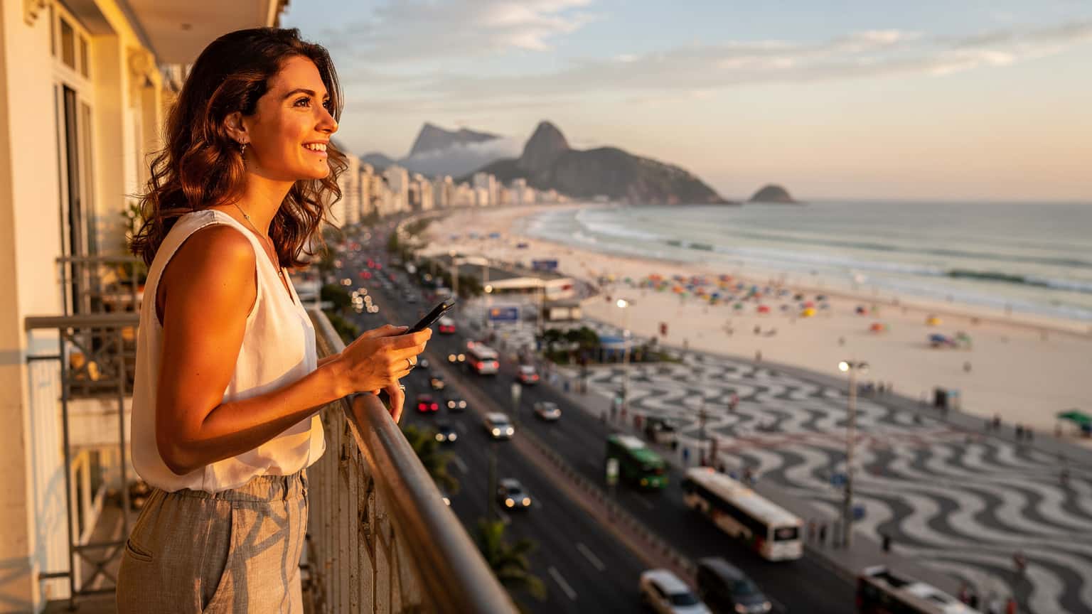Eine Frau mit einem Smartphone in der Hand, lächelt und blickt über die belebte Copacabana in Rio de Janeiro, während die Sonne untergeht.